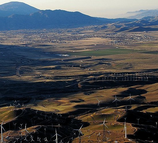 Aerial view over the Tehachapi Pass wind farm at golden hour — turbines on the ridgelines with the Tehachapi Valley and mountains beyond