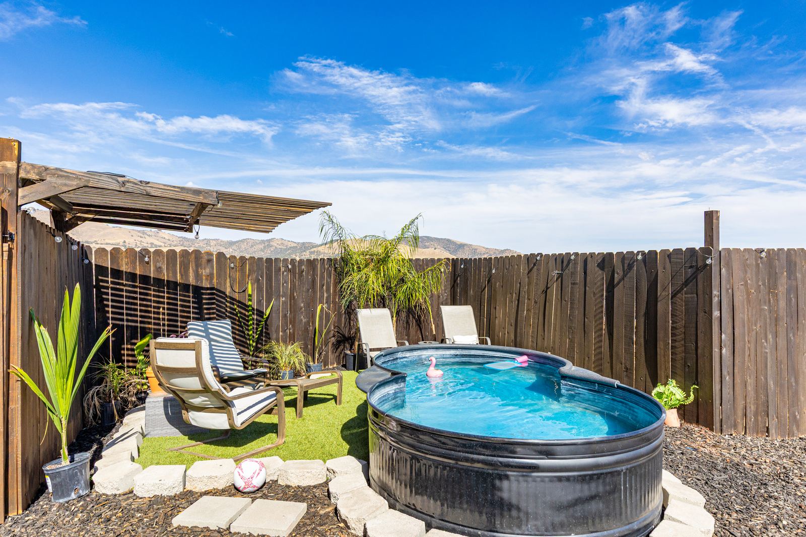 Fenced backyard corner with stock-tank pool, turf lounge area and hillside views